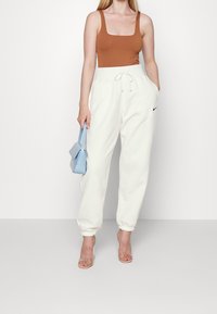 Žena v hnědém tílku, bílých teplákách Nike, průhledných lodičkách na vysokém podpatku a držící malé modré psan íčko.