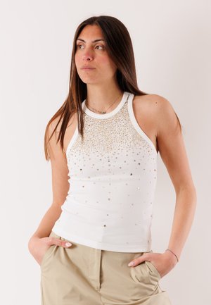 Donna che indossa una canotta bianca con borchie dorate, pantaloni beige, mani nelle tasche, che guarda leggermente di lato su uno sfondo neutro.