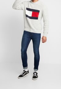 Grau strukturierter Pullover mit einem marineblauen, weißen und roten Logo auf der Brust, kombiniert mit dunkelblauen Slim-Fit-Jeans und schwarzen Turnschuhen.