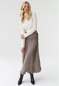 Pitkät, laskostetut hameet taupe-väristä kangasta, yhdistettynä istuvaan, kermaiseen turtleneck-paitaan. Mallilla on jalassaan mustat nilkkurit, joissa on sileät pinnat ja yksinkertainen muotoilu.