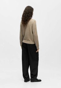 Une personne avec de longs cheveux bouclés se tient de dos, portant un pull beige, un pantalon noir à jambes larges et des chaussures noires sur un fond uni.