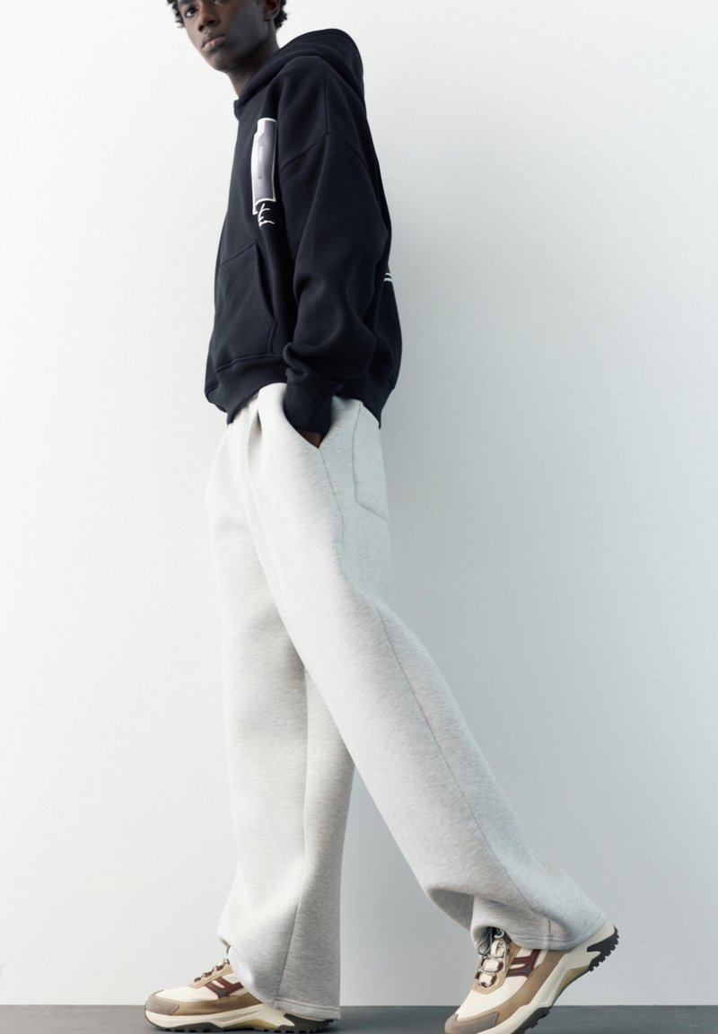 Jeune homme portant un sweat à capuche noir, un pantalon large gris clair et des baskets beiges, debout de profil sur un fond blanc uni.