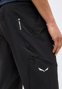 Κοντινό πλάνο ενός ατόμου που φοράει μαύρο παντελόνι Durastretch με φερμουάρ στην τσέπη και λευκό λογότυπο στον μηρό, το χέρι ακουμπάει δίπλα από το πόδι.