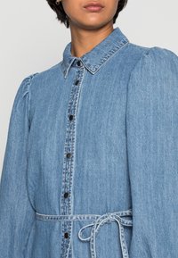 Jeansklänning i ljusblått med krage, knappstängning fram och puffärmar. Har ett knytliv och kontrasterande sömdetaljer.