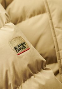 Chaqueta puffer en color marrón con superficie texturizada y costuras visibles. La etiqueta de la marca está bordada en negro, rojo y beige.