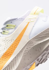 Chaussure de course légère avec une tige crème texturée ornée d'accents orange et jaune, des panneaux en mesh, et une semelle à motifs pour une meilleure adhérence.