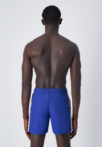 Pantaloni da bagno blu con cintura elastica. Presentano strisce laterali a contrasto in rosso e bianco. Tessuto leggero, design fino al ginocchio.