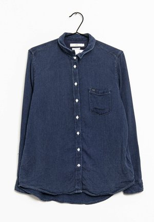 Camicia in denim blu scuro testurizzata a maniche lunghe con bottoni bianchi e taschino sul petto appesa a una gruccia nera su sfondo bianco.