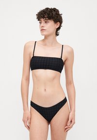 Crni bikini s bandeau topom s tankim trakama, teksturiranim uzorkom i odgovarajućim donjim dijelom niskog struka. Glatka tkanina s minimalnim detaljima.