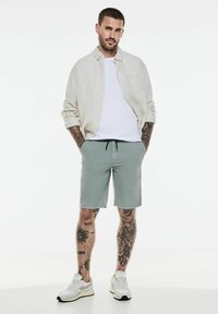 Chemise en lin beige clair, t-shirt blanc et shorts vert menthe. Baskets blanches avec des accents noirs. Tatouages visibles sur les jambes.
