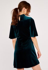 Abito in velluto teal con maniche corte e svasate e apertura a chiave sul retro. Tessuto morbido, vita aderente e orlo svasato.