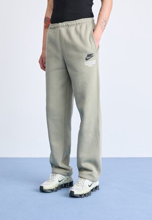 Person trägt hellgraue Nike-Sweatpants mit dem Text "Authorized Personnel Only" und weiße Nike-Sneaker und steht auf blauem Boden.