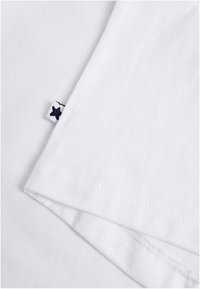 T-shirt en coton blanc avec une manche repliée, comportant une petite étiquette en forme d'étoile bleue cousue dans l'ourlet. Texture douce, design minimaliste.