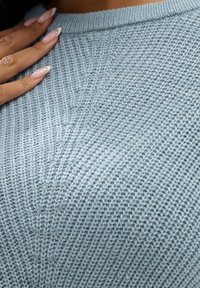 Suéter de punto azul claro con un patrón texturizado, cuello acanalado y vista en primer plano con los dedos descansando sobre la tela.