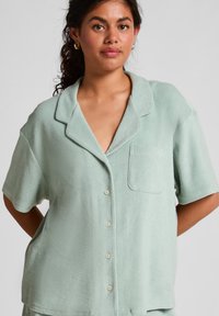 Jonge vrouw met krullend donker haar draagt een lichtgroen overhemd met korte mouwen en borstzak, staand tegen een effen achtergrond.