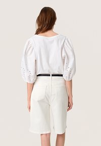 Hvid bomuldsbluse med puffede ærmer og broderede detaljer; parret med hvide denimshorts. Bagpå ses bæltestrop og lommeindretning.