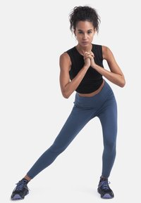 Musta hihaton toppi, siniset korkeavyötäröiset legginsit ja mustat urheilusandaalit purppuranvärisillä yksityiskohdilla. Malli on dynaamisessa asennossa.