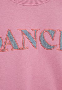 Roza tkanina z našitimi črkami, ki imajo teksturo jeansa in koralno nitjo za obrobo, ki tvorijo del besede z velikimi črkami.