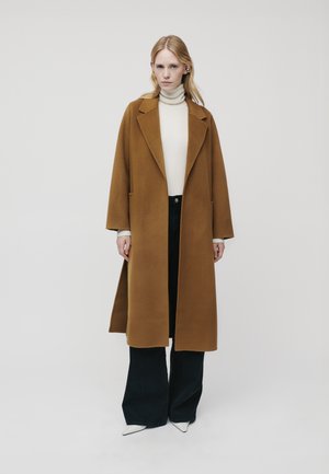 Cappotto di lana marrone con colletto a risvolto, vestibilità oversize e tasche laterali, abbinato a un dolcevita crema e jeans svasati scuri, completato da scarpe bianche.