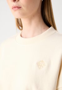 Sweat-shirt couleur crème avec un col côtelé. Présente un logo brodé sur la poitrine. Texture douce, design décontracté.