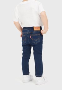Mörkblå denimjeans med smal passform, med en ljusbrun läderpatch baktill, gula sömmar och två bakfickor. Uttagen med en vit T-shirt.