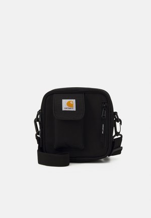 Sac bandoulière noir Carhartt avec poche avant à rabat, poche zippée, sangle réglable et patch logo sur fond clair.