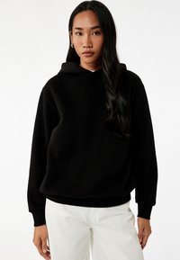Schwarzer Kapuzenpullover mit lockerer Passform, weichem Material und gerippten Bündchen. Kombiniert mit hellen Hosen für den Kontrast.