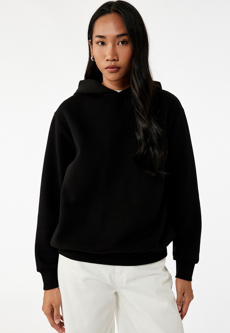 Schwarzer Kapuzenpullover mit lockerer Passform, weichem Material und gerippten Bündchen. Kombiniert mit hellen Hosen für den Kontrast.