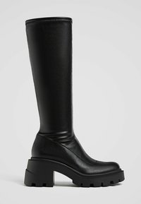 Bottes noires montantes avec un talon épais et une semelle crantée, en cuir lisse avec un bout rond, présentées en profil latéral sur fond blanc.