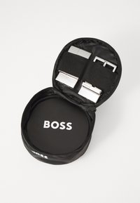 Étui zippé noir contenant deux boucles de ceinture en métal argenté maintenues par des sangles noires, avec une base circulaire portant le nom de la marque "BOSS."