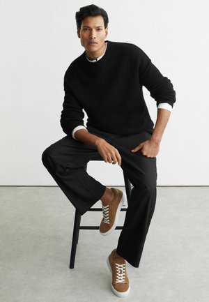 Mees istumas mustal pinkil, kandes musta kampsunit valge särgi üle, musti pükse ja pruune tossusid valgete paeltega hallil põrandal.