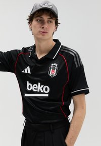 Juoda trumpomis rankovėmis polo marškinėliai su raudonomis detalėmis, balta apykakle ir siuvinėta logotipu. Derinami su juodomis aukšto liemens kelnėmis ir pilka kepure.