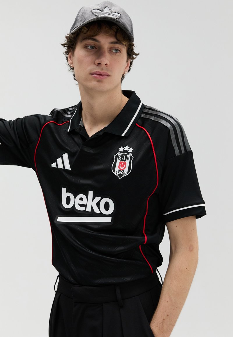 Juoda trumpomis rankovėmis polo marškinėliai su raudonomis detalėmis, balta apykakle ir siuvinėta logotipu. Derinami su juodomis aukšto liemens kelnėmis ir pilka kepure.