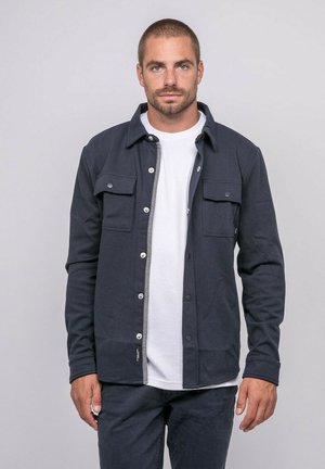 Homme aux cheveux courts et barbe portant une veste boutonnée gris foncé par-dessus un t-shirt blanc, debout devant un fond clair uni.