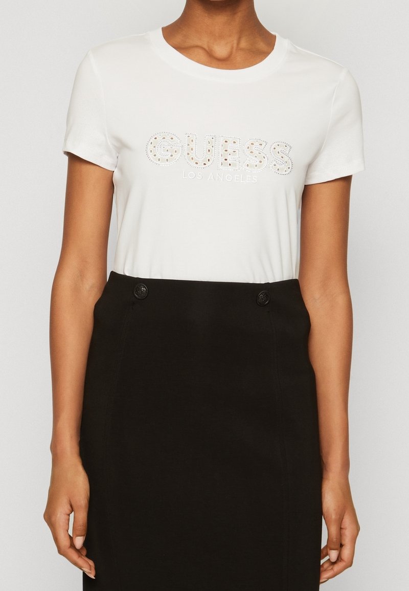 Femme portant un t-shirt blanc à manches courtes avec le texte "GUESS LOS ANGELES" associé à une jupe noire taille haute avec des boutons.