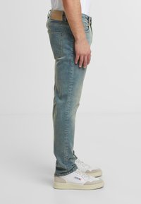 Světle modré džíny s úzkým střihem, se zadní kapsou a hnědou koženou nášivkou na pase. Nosené s bílými teniskami.