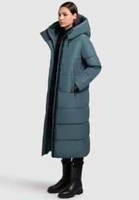 Długa puchowa kurtka w kolorze morskim z wysokim kołnierzem, zapięciami na zamek i guziki, bocznymi kieszeniami oraz pikowanym wzorem. Noszona z czarnymi butami i legginsami.