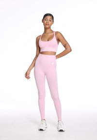 Svetlo roza športni modrček s tankimi naramnicami, v kombinaciji z ujemajočimi se visokimi pajkicami. Ima majhen bel logotip na pajkicah.