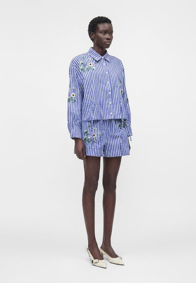 Femme portant une chemise à rayures verticales bleues et blanches à boutons et un short assorti avec une broderie florale, associée à des talons pointus blancs.