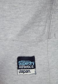 Un tessuto grigio con una superficie liscia presenta un'etichetta blu e bianca "Superdry Athletics Japan" cucita su di esso.