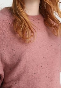 Różowy sweter z okrągłym dekoltem, wykonany z teksturowanej tkaniny, z małymi ciemnymi plamkami na całej powierzchni. Miły i przytulny wygląd.