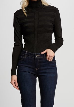 Femme portant un pull à col roulé côtelé noir avec des détails de rayures horizontales et un jean skinny bleu foncé sur un fond uni.