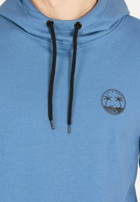 Blå hoodie med funnel neck, svarta snören och en cirkulär tryckt logotyp med palmträd och vågor. Mjuk, texturerad tyg.