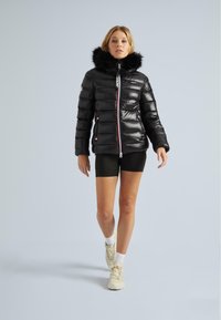 Žena jde a má na sobě černou péřovou bundu s kožešinovou kapucí, černé šortky, bílé ponožky a béžové tenisky na jednolitém světle šedém pozadí.