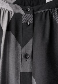 Tissu à motifs noir et gris avec une patte de boutonnage comportant deux boutons noirs. La texture générale est lisse avec un léger éclat.