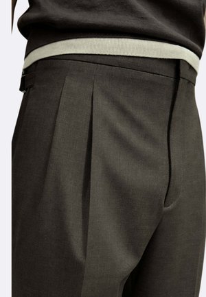 Primo piano di pantaloni sartoriali grigio scuro con pieghe, indossati con una maglia nera infilata nella vita e rifinita con una bordatura grigio chiaro.