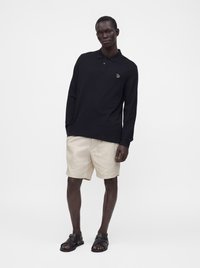 Homme portant un polo noir à manches longues, un short beige et des sandales noires, debout devant un fond blanc uni.