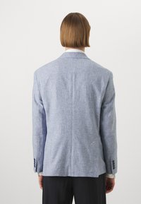 Gaišu zilu lina žaketi ar teksturētu apdari, ar izgrieztiem laciem, vienu ventiļu un trīs pogām aprocēs, kopā ar melniem biksēm.