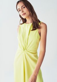 Rankovi bešvarkiai, iki kelių ilgio suknelė iš ryškiai geltonos medžiagos. Pasižymi suknele susukta mazgo detale juosmens srityje ir sklandžiu, minimalistiniu dizainu.