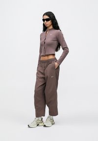 Μπλούζα με μακριά μανίκια, ριμπ ύφασμα και κουμπιά σε μωβ απόχρωση, συνδυασμένη με φαρδιά παντελόνια με ελαστική μέση σε σκουρότερο καφέ χρώμα, και τα δύο με λογότυπο Nike.